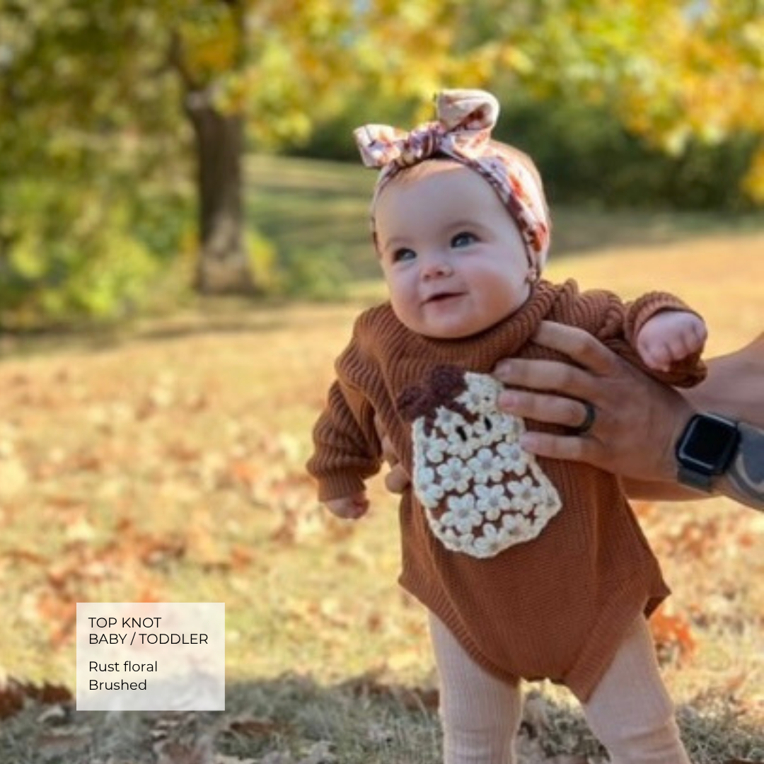 A baby wearing a rust brown knit top, cream knit belly patch, and a floral-patterned top knot headband, held outdoors by an adult's hand. A text box in the bottom left reads 'TOP KNOT BABY / TODDLER Rust floral Brushed'.