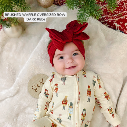 Baby wearing nutcracker print pajamas and a red brushed waffle oversized bow, under the Christmas tree.