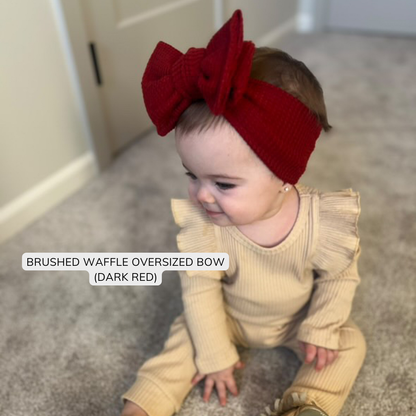 Baby wearing a dark red oversized bow headband and matching outfit on a neutral background