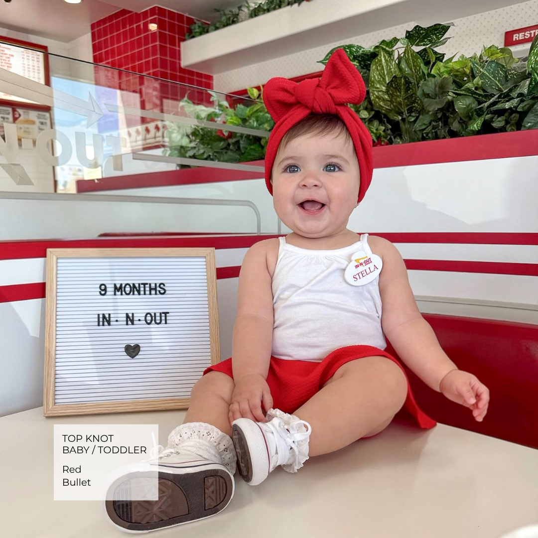 A baby sitting in a restaurant or cafe setting wearing a red top knot headband, white tank top, and red skirt. A letter board next to the baby reads '9 MONTHS IN-N-OUT' and a text overlay on the bottom left says 'TOP KNOT BABY/TODDLER Red Bullet'.