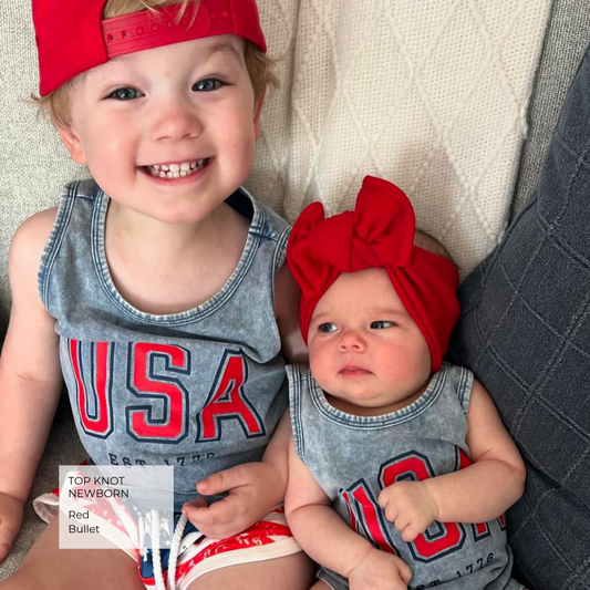 Two children wearing matching red and gray denim outfits with 'USA' lettering, posed together on a textured cream couch. The older child wears a red cap backward, and a watermark in the lower left reads 'TOP KNOT, NEWBORN Red Bullet.