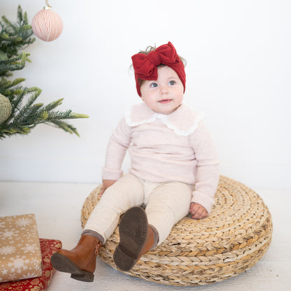 Baby sitting on a woven stool wearing a pink outfit and red headband, with Christmas decorations in the background.