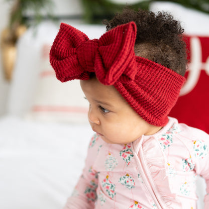 Baby wearing a red knitted headband with a large bow, sitting in front of a blurred background.