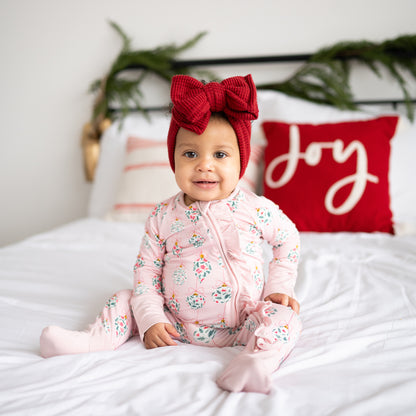 Baby in pink floral outfit with red headband sitting on a bed with Christmas decorations.