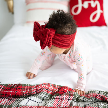 Baby wearing a red headband with a bow, sitting on a bed with Christmas-themed pillows.
