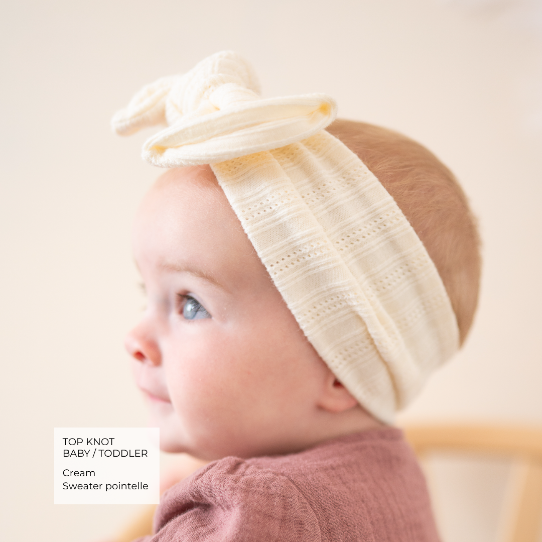 A baby wearing a cream knit headband with a large top knot bow, shown in profile against a soft beige background. A text box in the lower left reads 'TOP KNOT BABY / TODDLER Cream Sweater pointelle'.