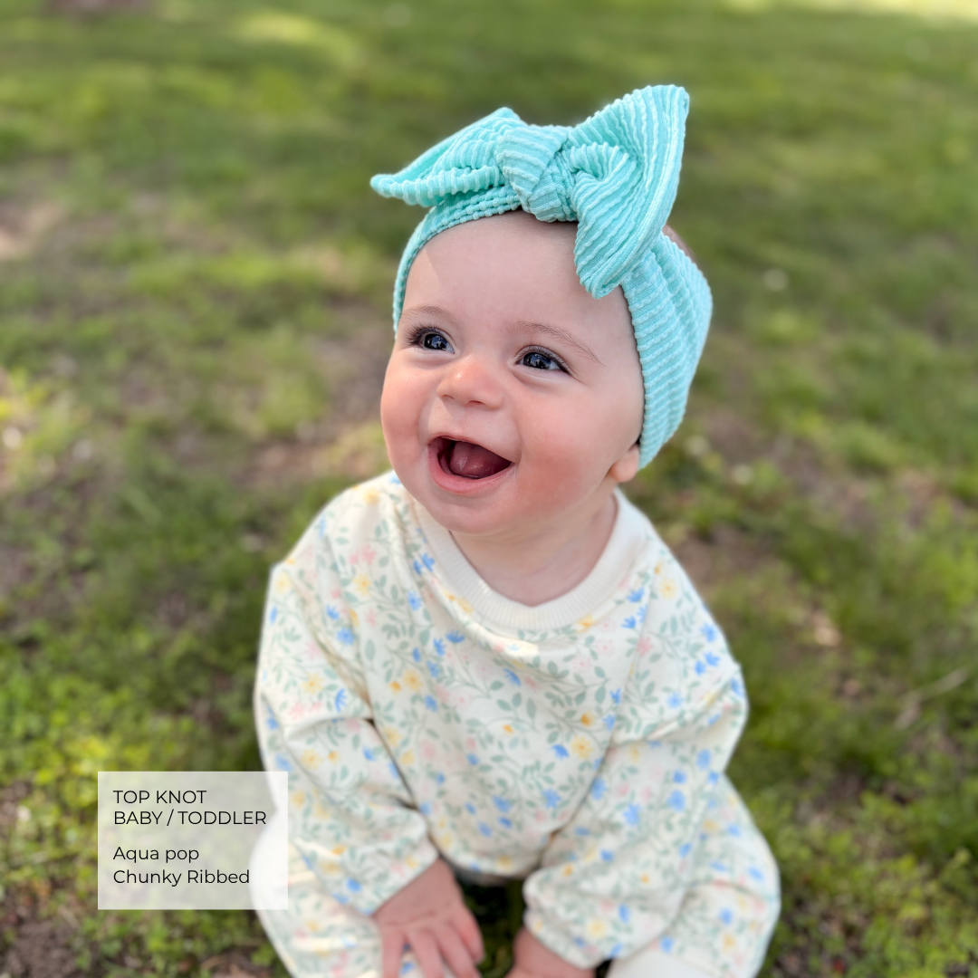 A smiling baby wearing a light aqua ribbed top knot headband and a cream floral long-sleeve romper, sitting outdoors on a grassy lawn. A text overlay in the bottom left reads 'TOP KNOT BABY/TODDLER Aqua pop Chunky Ribbed'.
