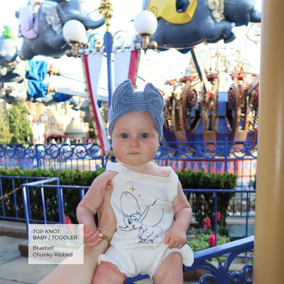 Baby wearing a blue chunky ribbed top knot and white onesie with a cartoon design, sitting in front of the Dumbo ride at Disneyland.