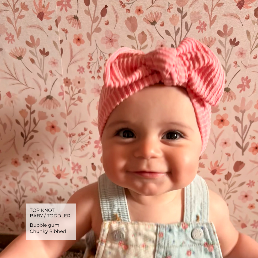 A baby wearing a pink chunky ribbed top knot headband and light blue denim overalls, set against a floral wallpaper background. A text box in the bottom left corner reads 'TOP KNOT BABY / TODDLER Bubble gum Chunky Ribbed'.