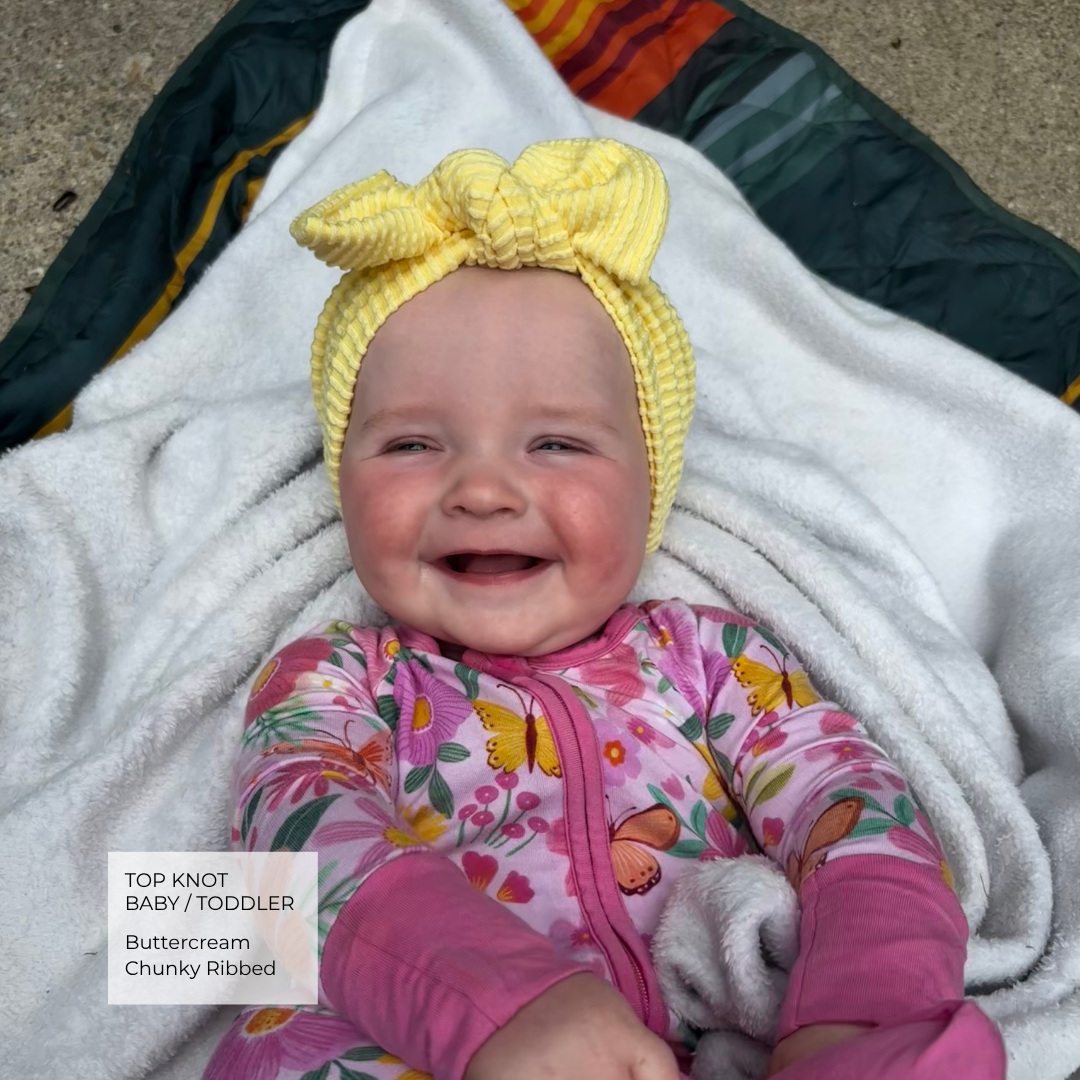 A baby wearing a yellow chunky knit top knot headband and a pink butterfly-print footed sleeper, lying on a white blanket outdoors.