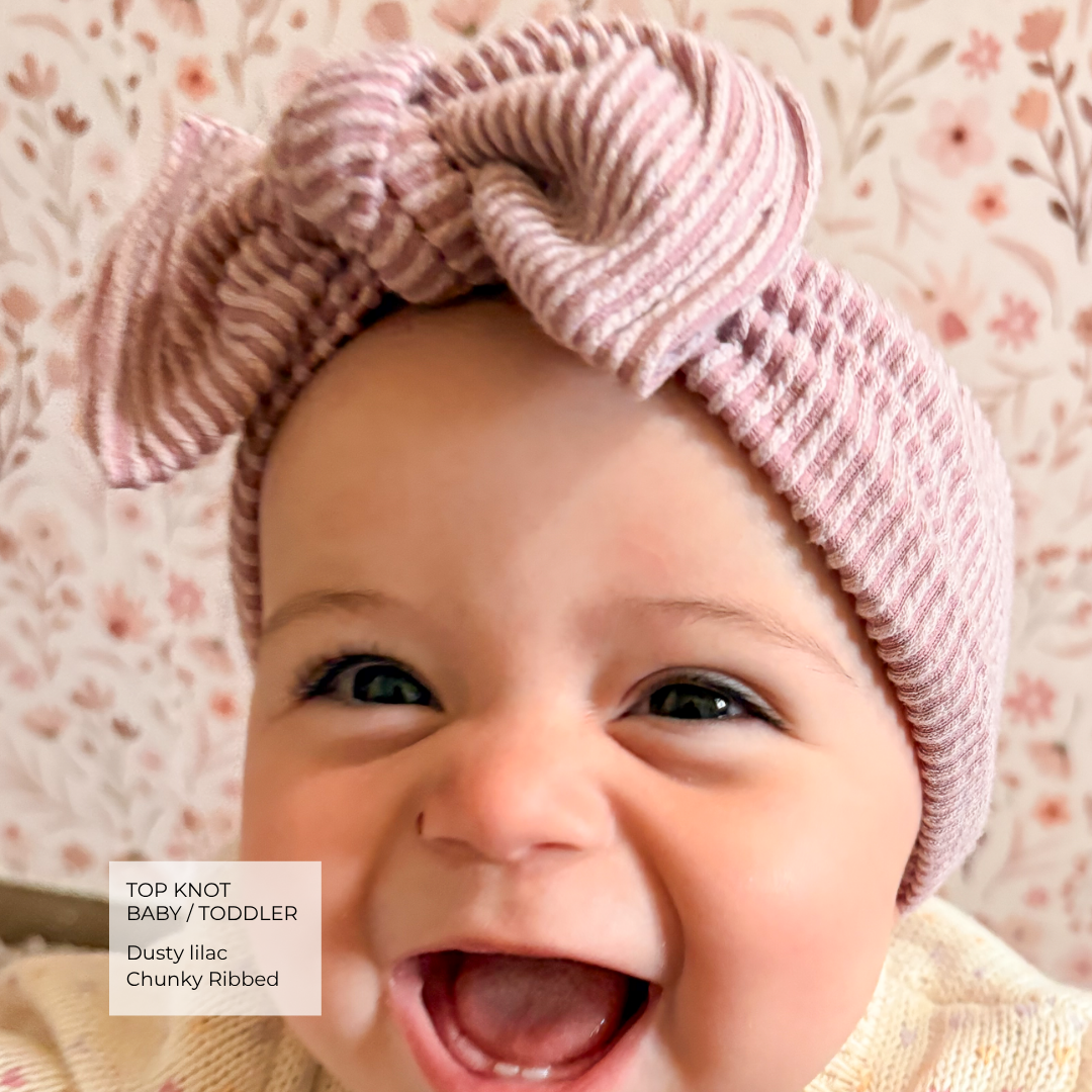 A close-up of a smiling baby wearing a chunky ribbed knit top knot headband in dusty lilac-pink, with a floral wallpaper background and a text label in the lower left corner.