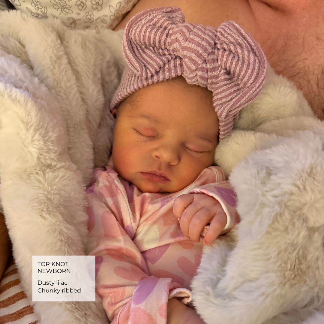 A sleeping newborn baby wearing a dusty lilac and pink striped top knot headband and matching patterned footie, nestled in a white faux fur blanket.