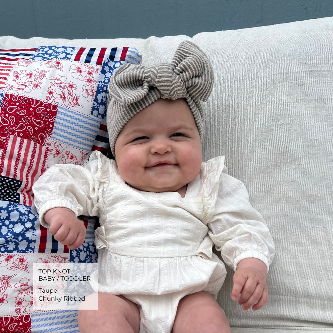 A baby wearing a chunky ribbed taupe top knot headband and a white long-sleeve romper, sitting on a white cushion next to a red, white, and blue patchwork pillow. A text overlay in the bottom left corner reads 'TOP KNOT BABY / TODDLER Taupe Chunky Ribbed'.