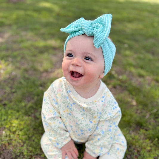 Baby wearing a turquoise knitted headband with a bow, sitting on grass.
