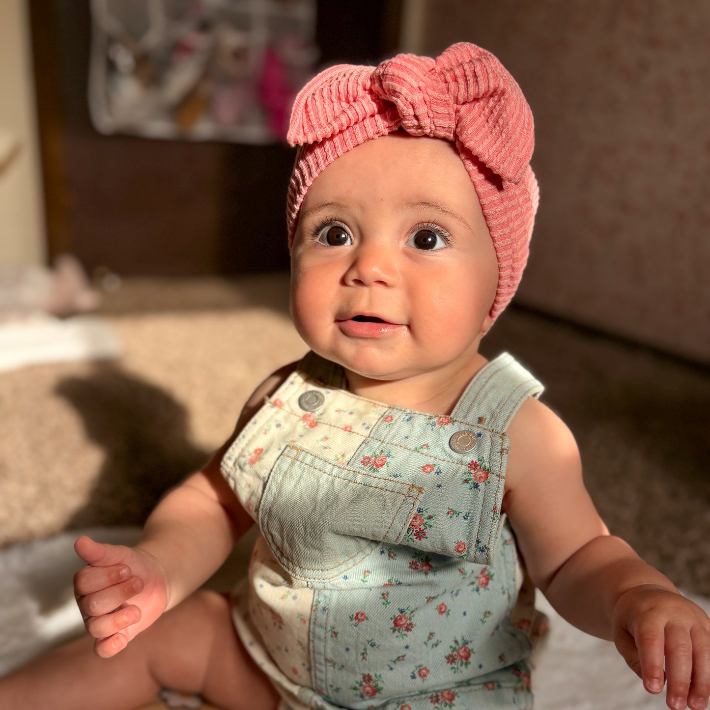 Baby wearing a pink headband and floral overalls sitting on a blanket.
