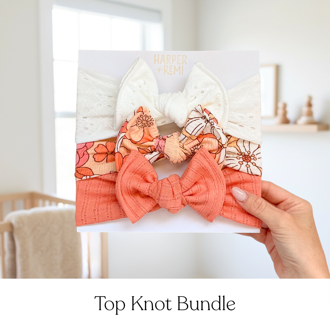 A baby hair bow set on a white display card, held by a hand in a softly lit nursery. The set includes a large white lace bow, a coral pink textured bow, and two floral elastic bands in orange, pink, and white.