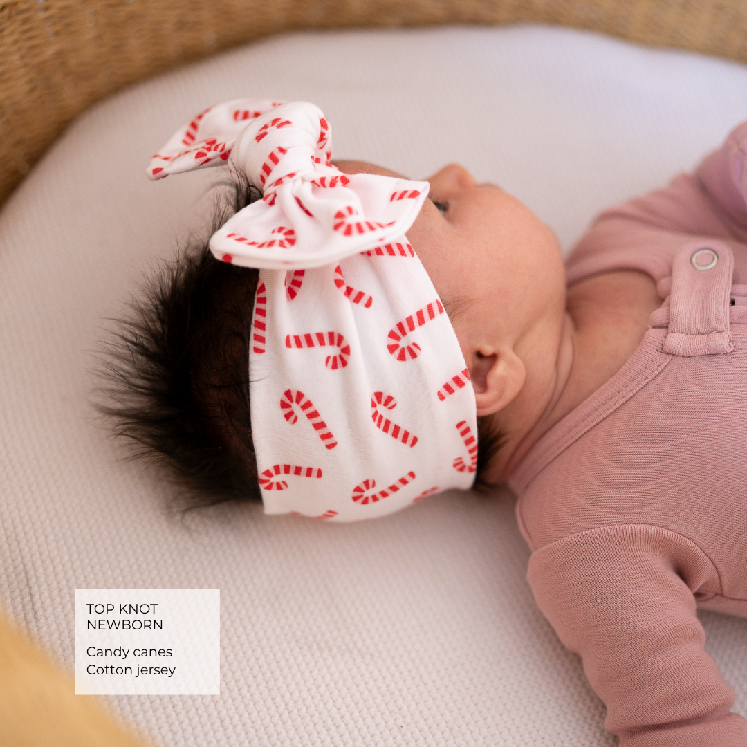 A newborn baby wearing a white cotton knit headband with a red candy cane pattern and a top knot, lying on a white blanket. The baby is wearing a dusty pink ribbed long-sleeve bodysuit.