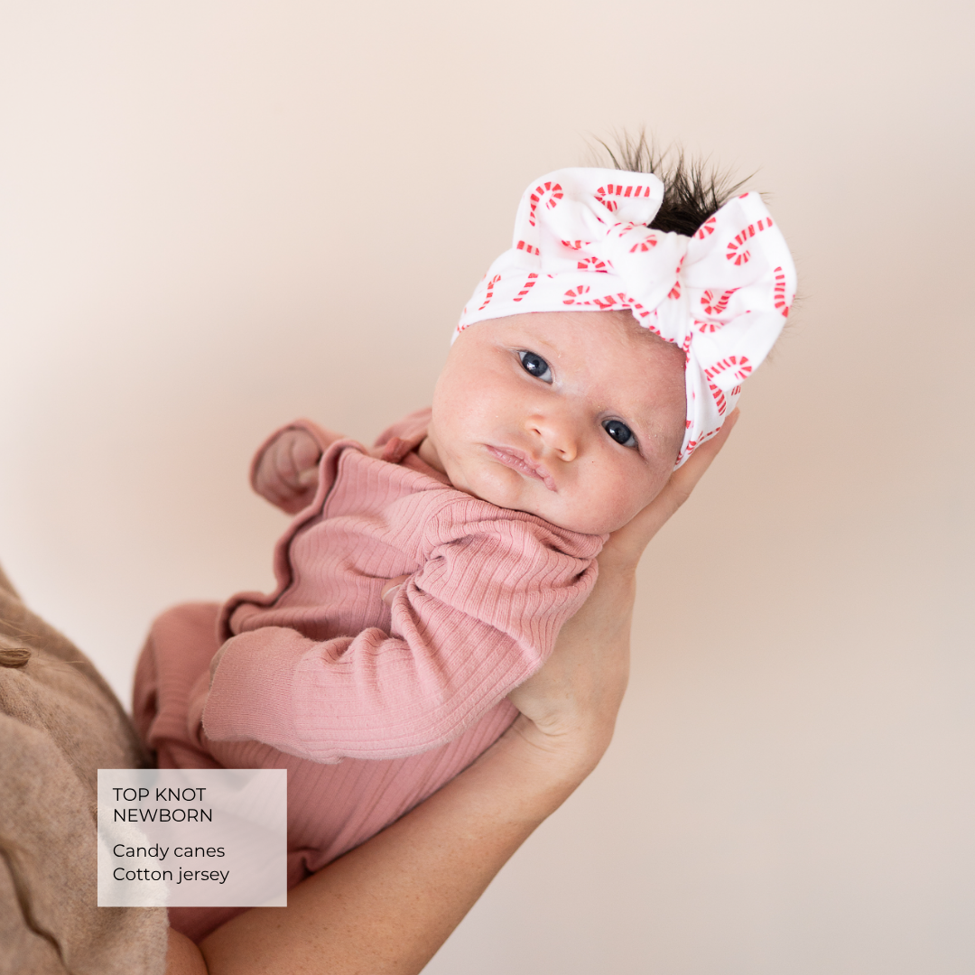 A newborn baby wearing a pink ribbed long-sleeve outfit and a white top knot headband with a red candy cane pattern, held in an adult's hands against a soft beige background. A text box in the bottom left reads 'TOP KNOT NEWBORN Candy canes Cotton jersey'.