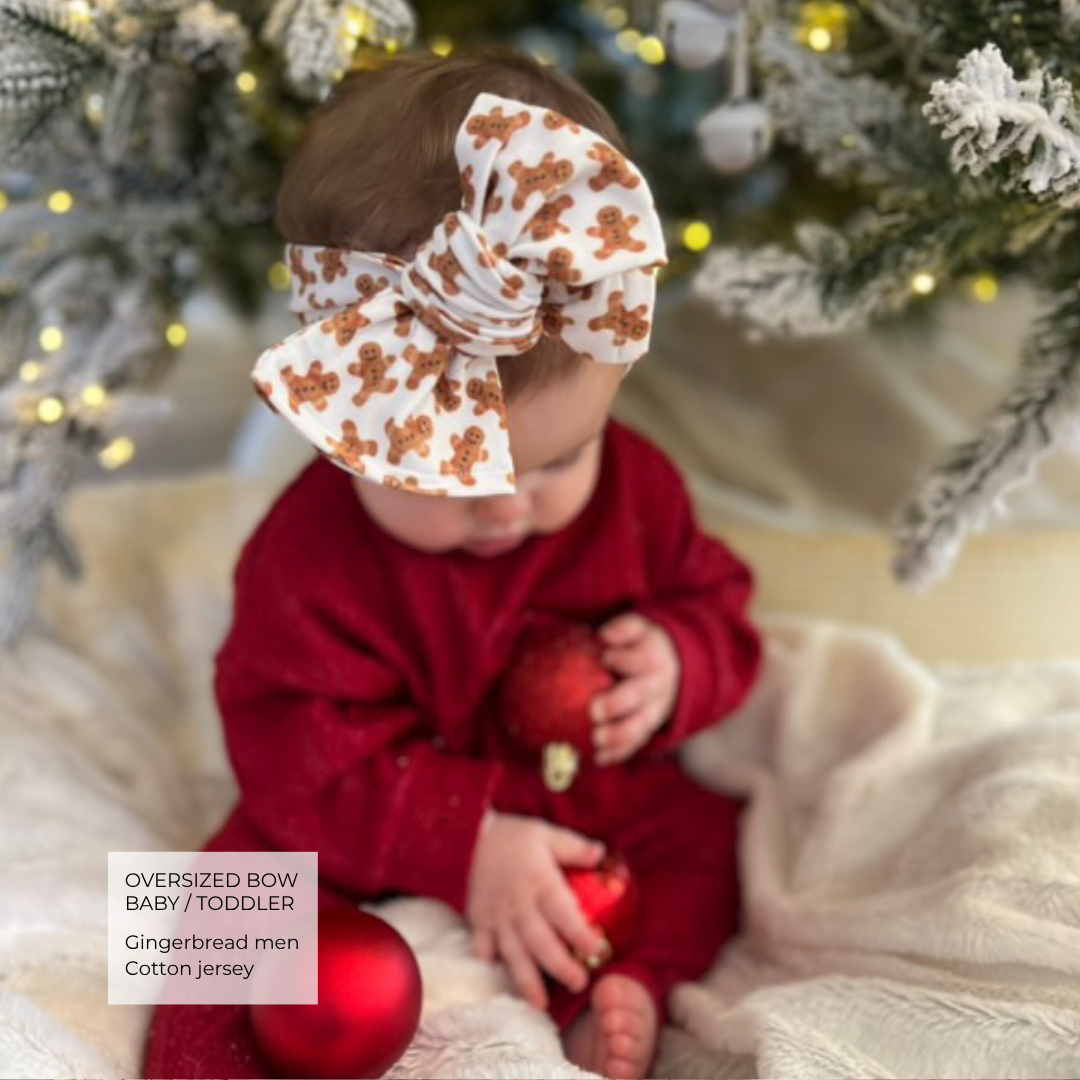 A baby wearing an oversized white cotton bow with a brown gingerbread man pattern, paired with a red jumpsuit, sitting in front of a decorated Christmas tree.
