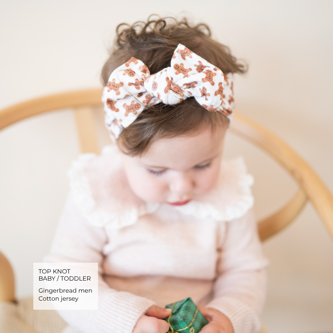 A toddler wearing a white cotton headband with a gingerbread man pattern, sitting in a wooden chair and looking down at a small green gift box.