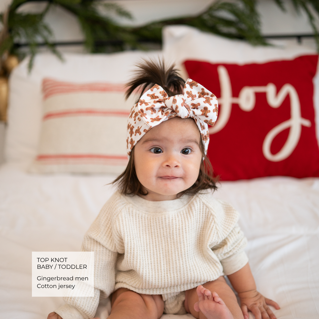 A baby toddler wearing a cream ribbed knit top and a matching cream top-knot headband with a gingerbread print, sitting on a white bed with a red 'Joy' pillow in the background.