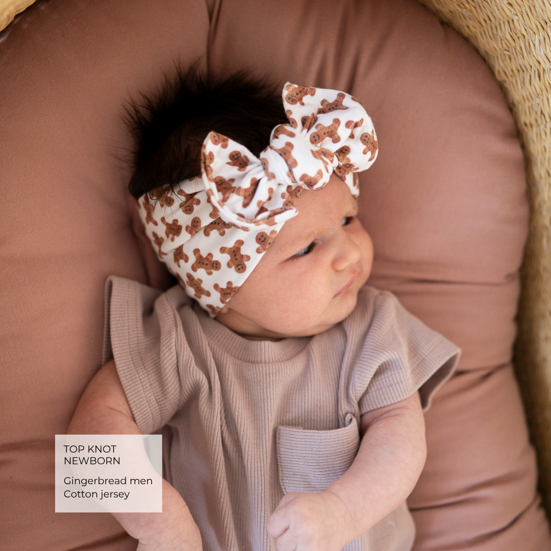 A newborn baby wearing a gingerbread men patterned top and a matching white top-knot headband with brown motifs, lying on a dusty rose cushion inside a wicker basket.