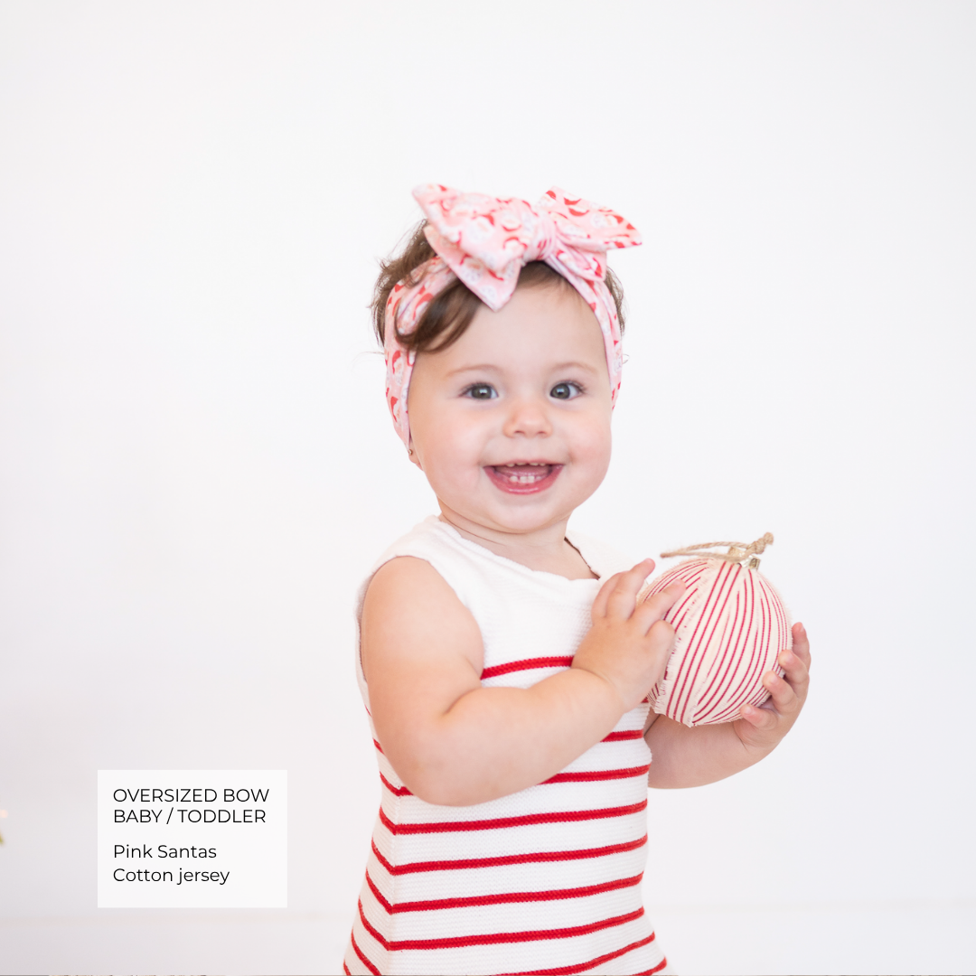 A smiling toddler wearing a pink and white striped cotton jersey outfit with a matching oversized bow headband, holding a small striped ball against a plain white background. Text in the lower left reads 'OVERSIZED BOW BABY / TODDLER Pink Santas Cotton jersey'.
