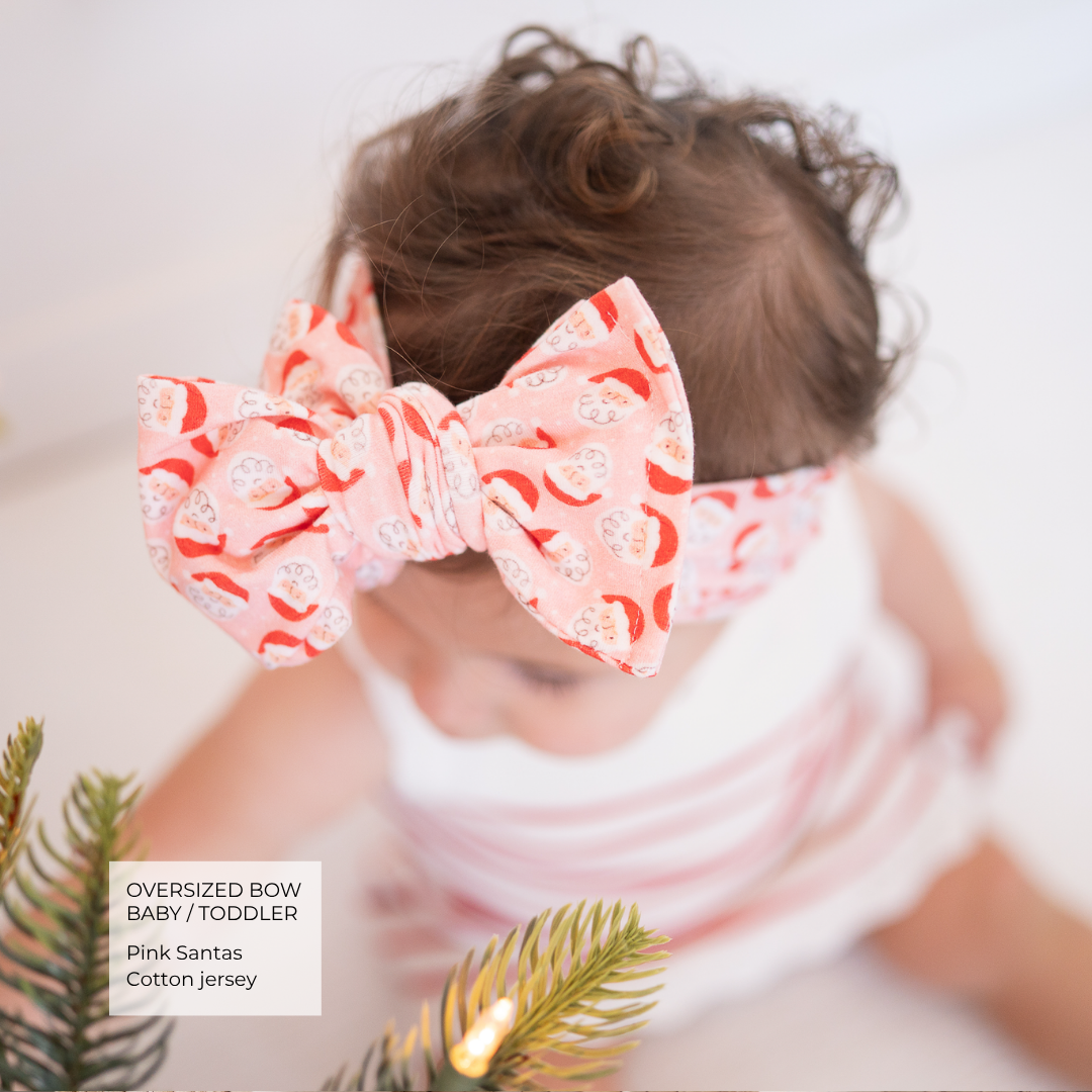 A toddler wearing a large oversized bow headband and matching headband in a pink cotton jersey fabric with a red Santa Claus pattern. The image features a soft-focus background and pine branches in the foreground, with a text overlay in the bottom left corner.