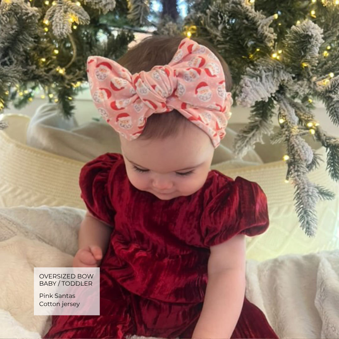 A toddler wearing an oversized pink Santa-patterned bow headband and a deep red velvet dress, sitting in front of a decorated Christmas tree. A text overlay in the bottom left reads 'Oversized Bow Baby/Toddler Pink Santas Cotton Jersey'.