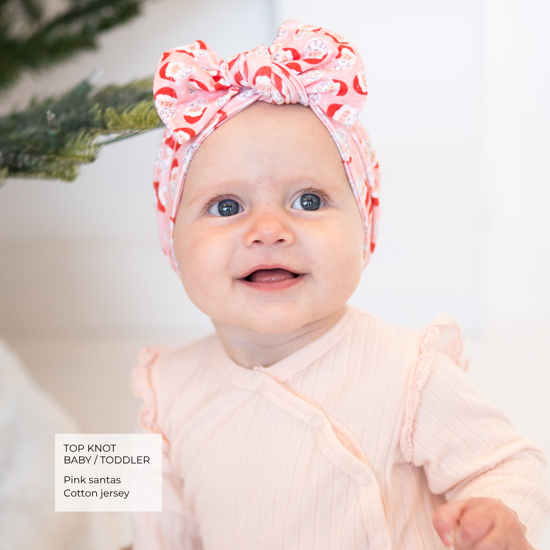 A smiling baby wearing a pink and red patterned top knot headband with a large bow, paired with a pale pink ribbed romper. A text box in the bottom left reads 'TOP KNOT BABY/TODDLER Pink santas Cotton jersey'.