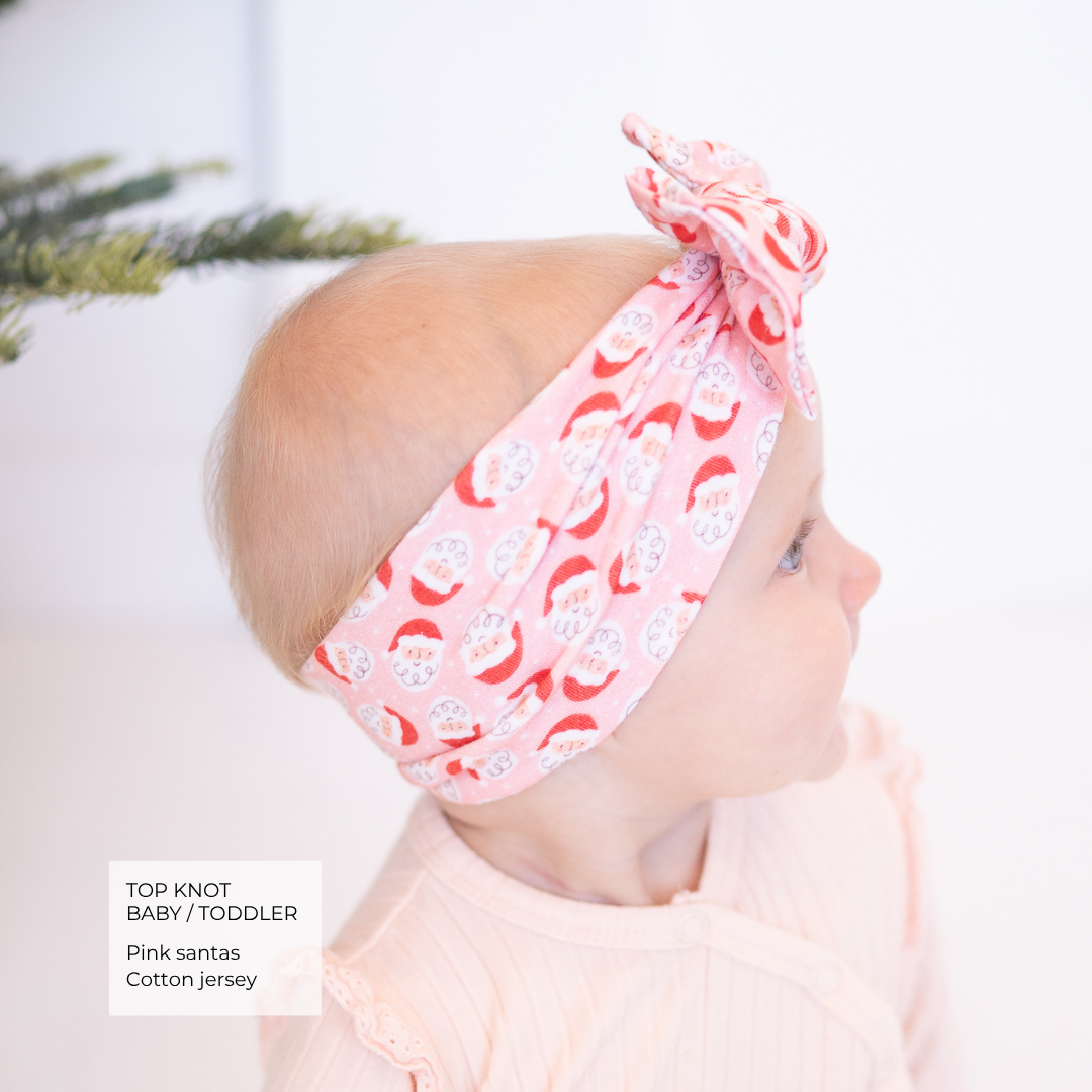 A baby in profile wearing a pink cotton jersey top and a matching pink Santa hat-top knot headband with a red and white Santa Claus pattern. A text box in the bottom left reads 'TOP KNOT BABY / TODDLER Pink santas Cotton jersey'.