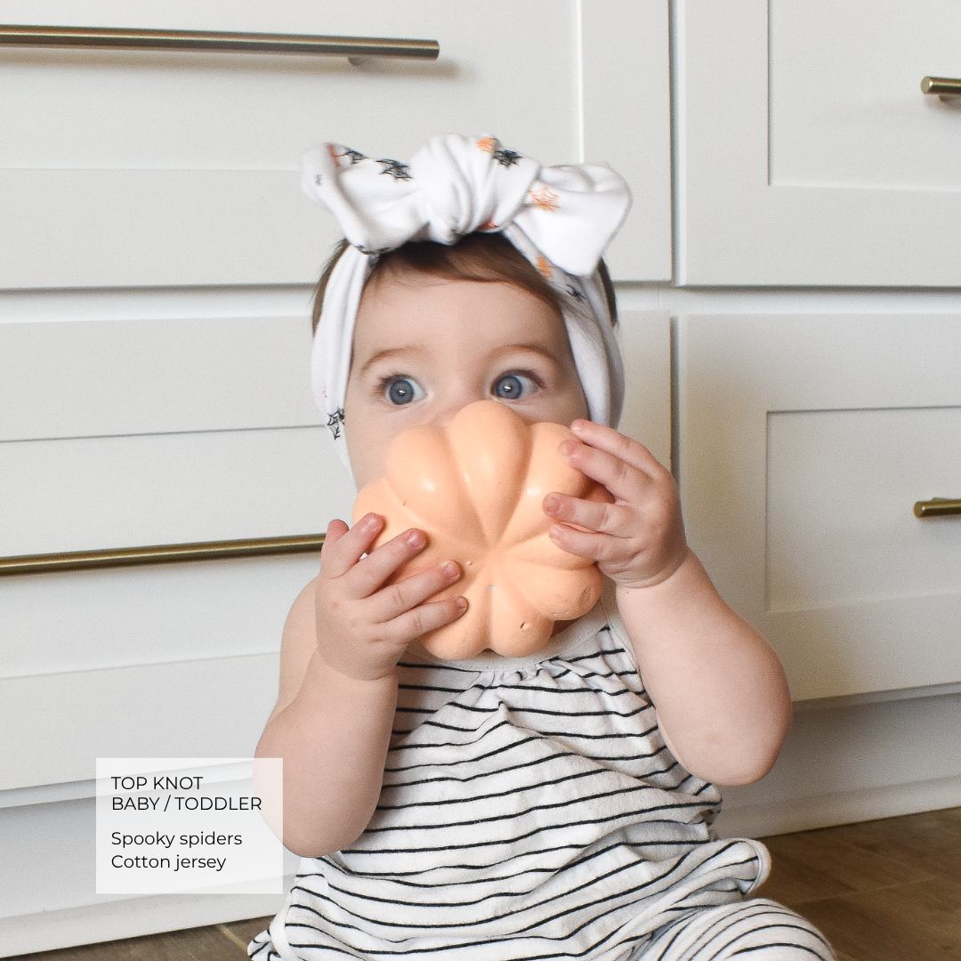 A baby wearing a white striped cotton jersey outfit and a white top knot headband with black spider prints, holding a peach-colored pumpkin toy in front of their face, set against a white kitchen cabinet background.