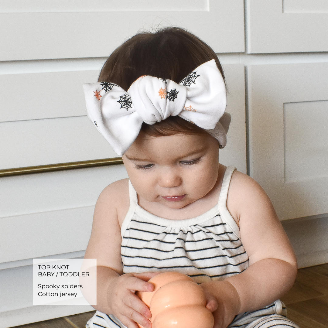 A toddler wearing a large white cotton jersey top knot headband with black spider and orange spiderweb prints, sitting in front of white cabinets and holding a peach-colored toy.