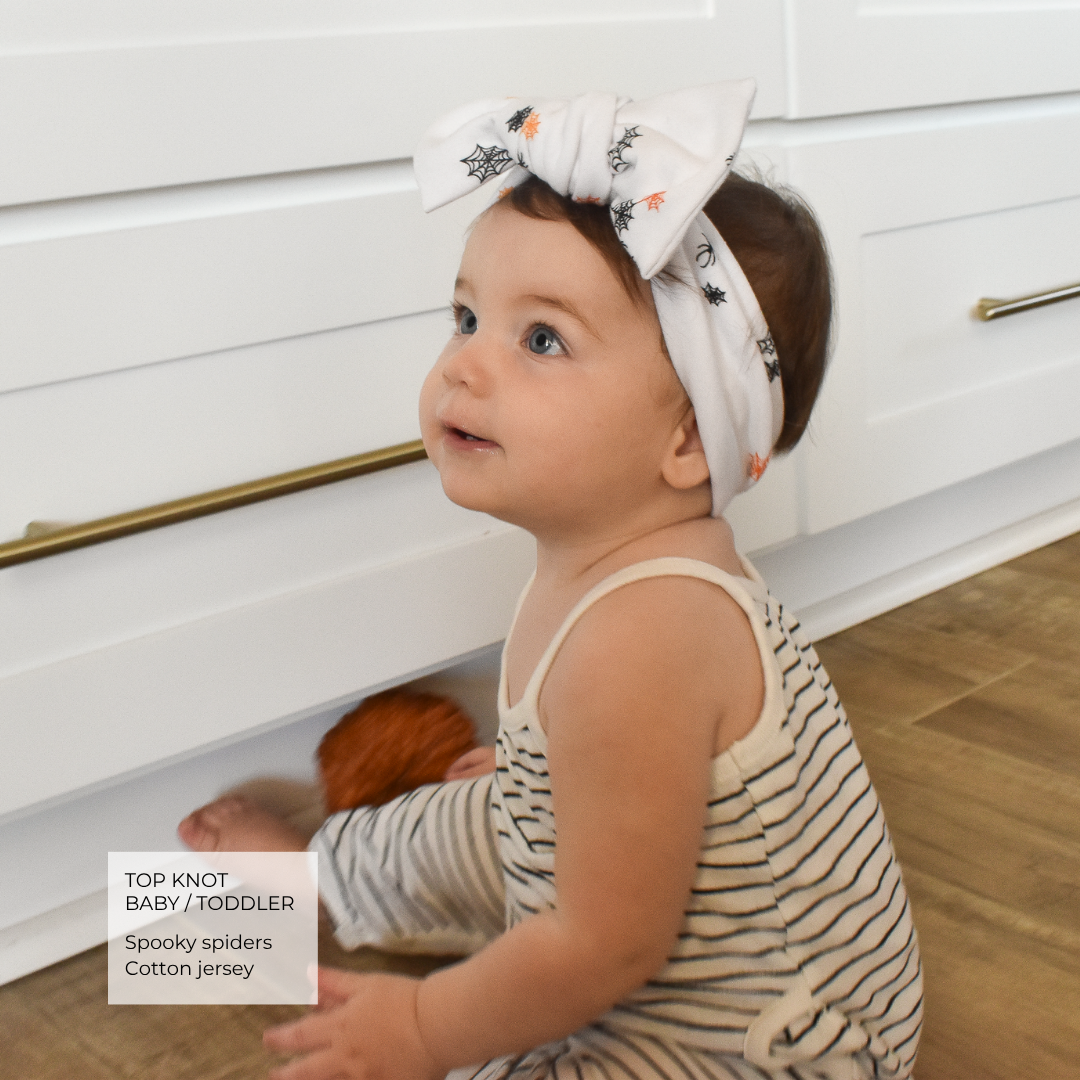 A baby wearing a white cotton jersey top and matching striped pants sits on a wooden floor in front of white cabinets, wearing a matching white top-knot headband with black spider and orange spiderweb prints. A text box in the bottom left reads 'TOP KNOT BABY / TODDLER Spooky spiders Cotton jersey'.