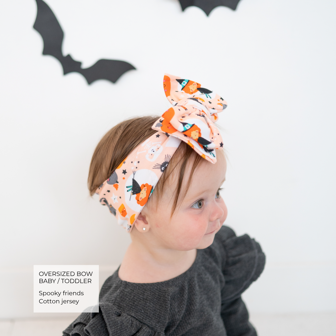 A toddler wearing an oversized fabric bow headband with a Halloween print of orange pumpkins, black bats, and white ghosts on a peach background. The toddler is wearing a dark gray long-sleeve top against a white wall with black bat decorations.