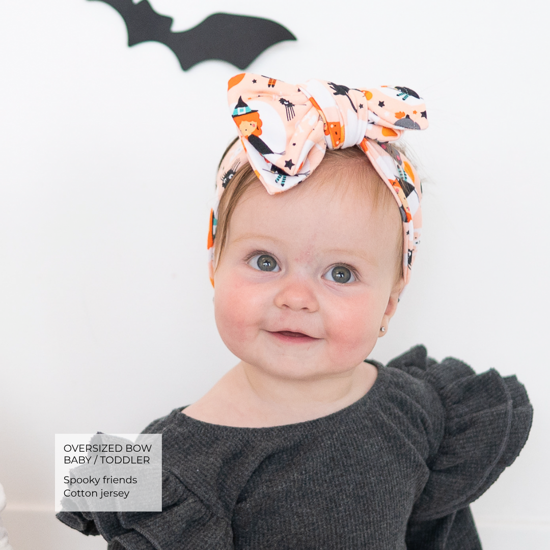 A baby wearing an oversized fabric bow headband with a Halloween print of orange pumpkins, black cats, and white stars on a peach background, paired with a dark gray ruffled top. A text overlay in the bottom left reads 'Oversized Bow Baby/Toddler Spooky friends Cotton jersey'.