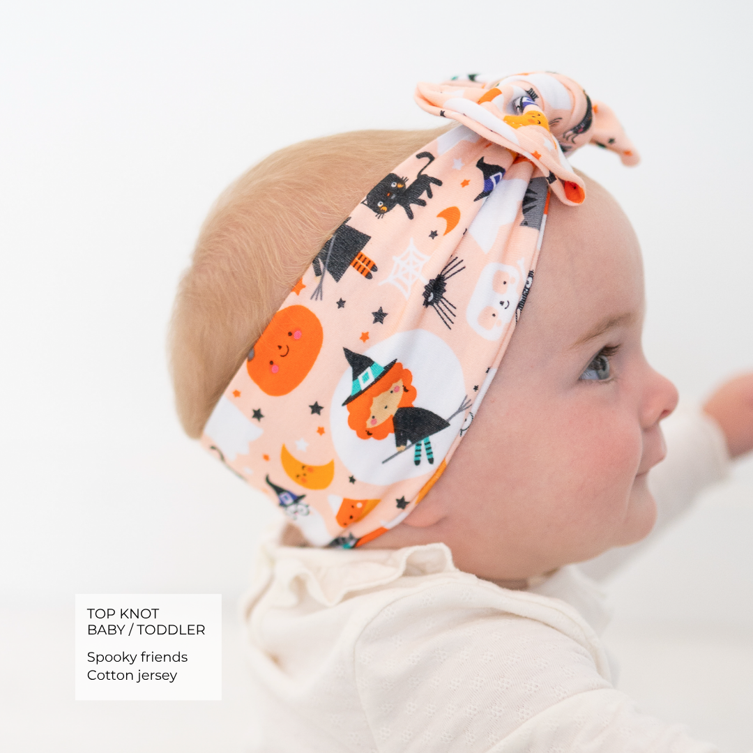 A baby wearing a cotton jersey top and a matching top knot headband with a spooky Halloween print, shown in profile against a white background. A text box in the bottom left lists the product details: 'TOP KNOT BABY / TODDLER, Spooky friends, Cotton jersey'.