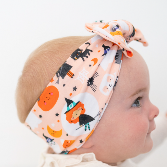A baby wearing a cotton jersey top and a matching top knot headband with a spooky Halloween print, shown in profile against a white background. A text box in the bottom left lists the product details: 'TOP KNOT BABY / TODDLER, Spooky friends, Cotton jersey'.