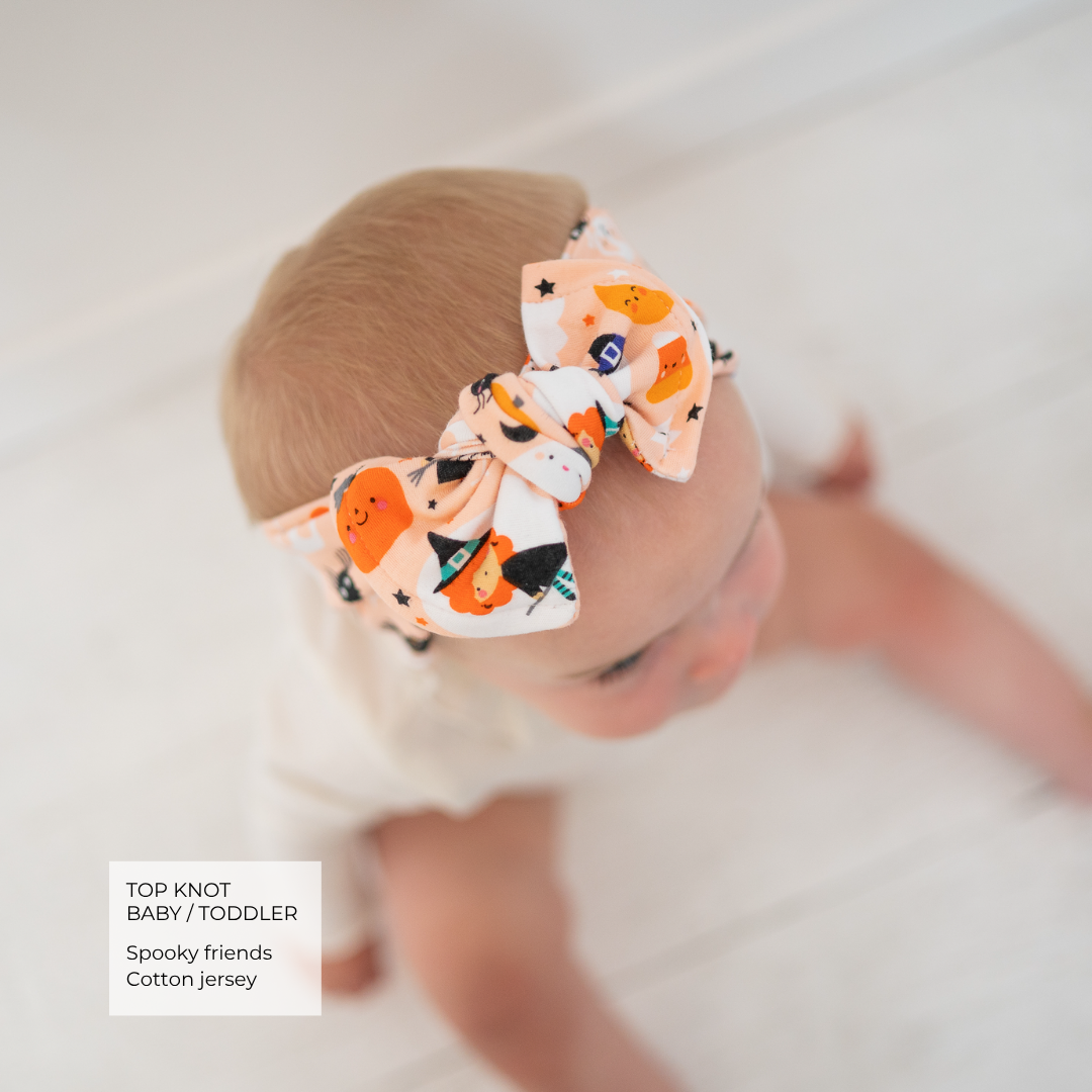 A baby wearing a large fabric bow headband with a Halloween print of orange pumpkins, black witches hats, and black stars on a white background. The text overlay in the bottom left reads 'TOP KNOT BABY / TODDLER Spooky friends Cotton jersey'.