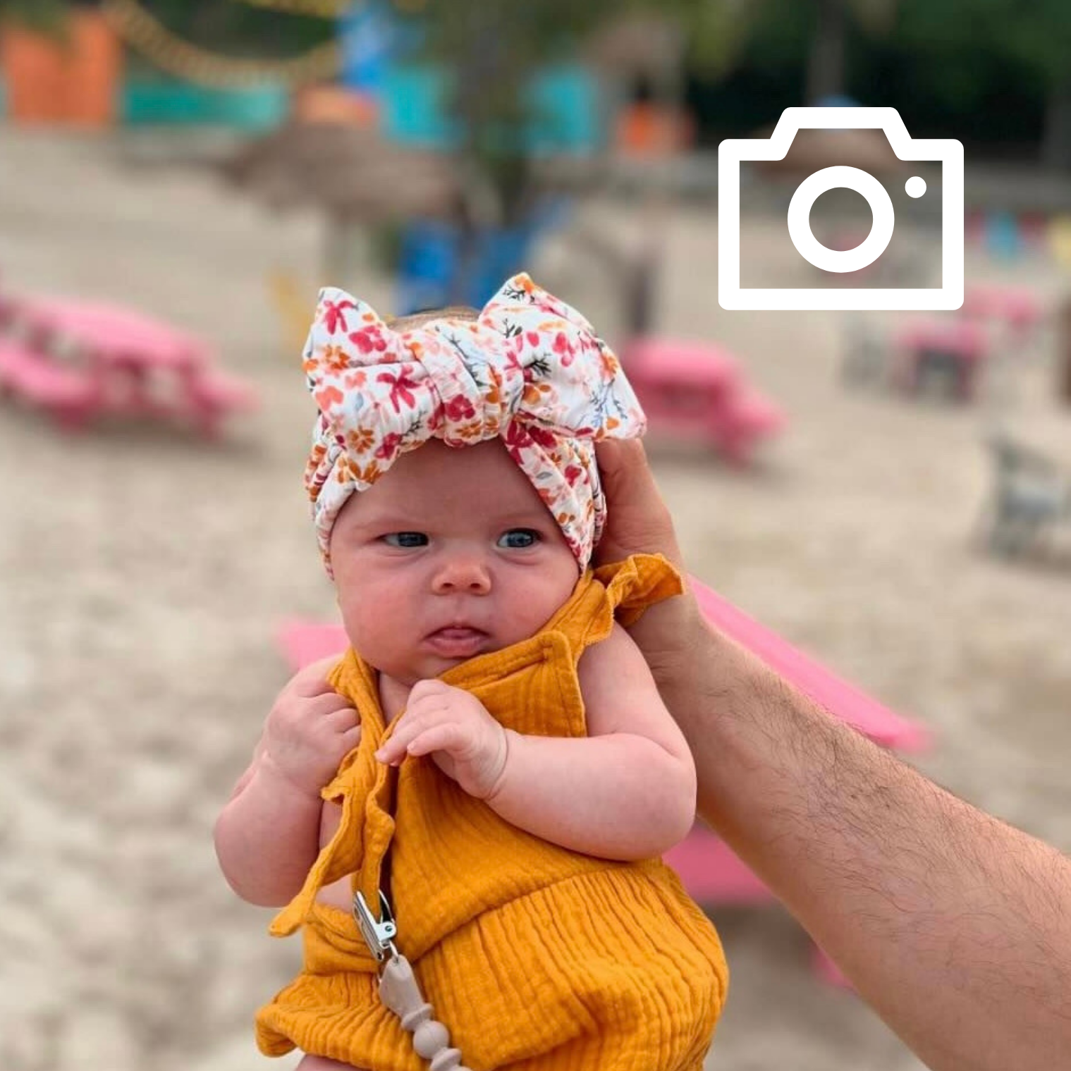 Baby in a yellow outfit with a floral headband at a beach.