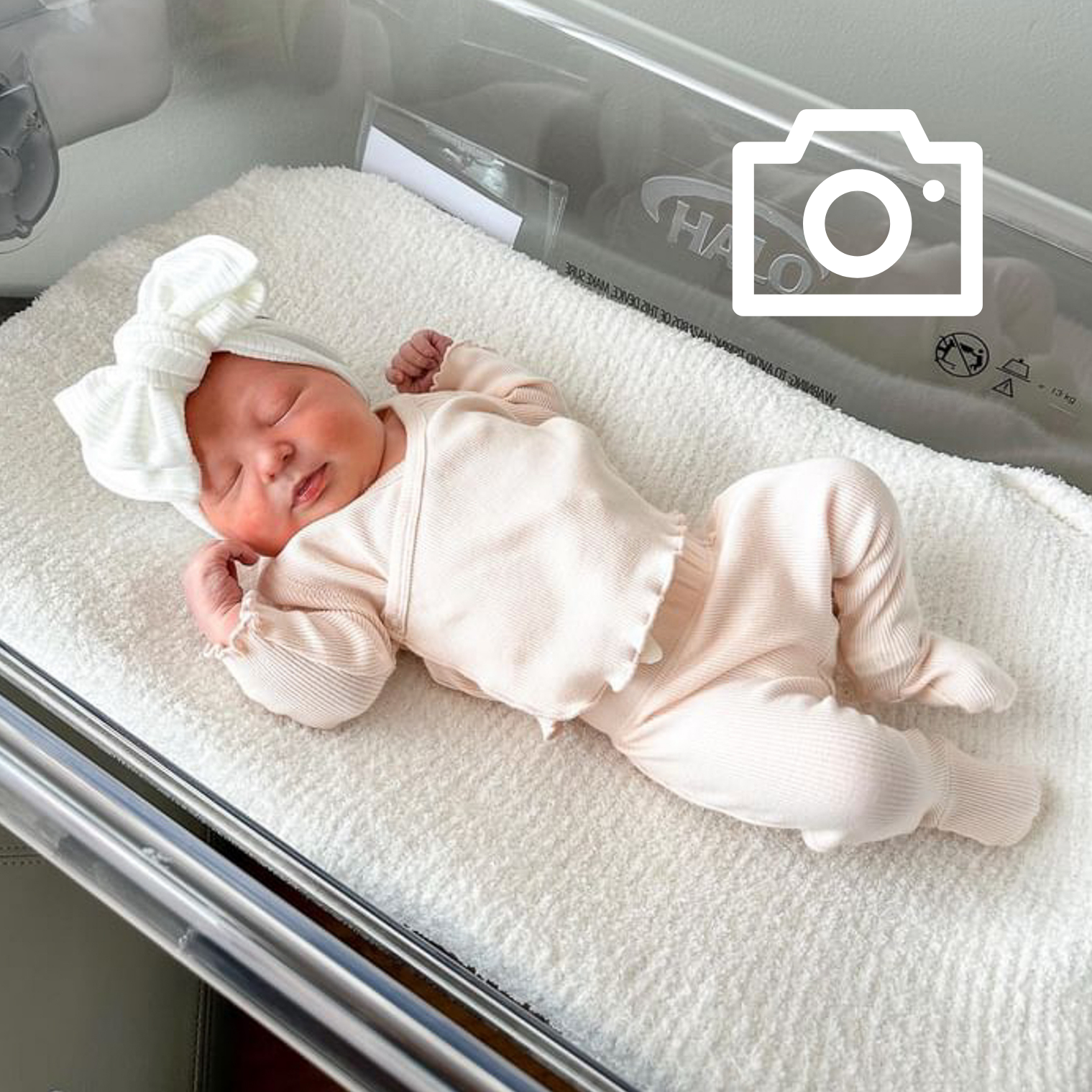 Newborn baby in a white outfit and hat lying on a hospital bed.