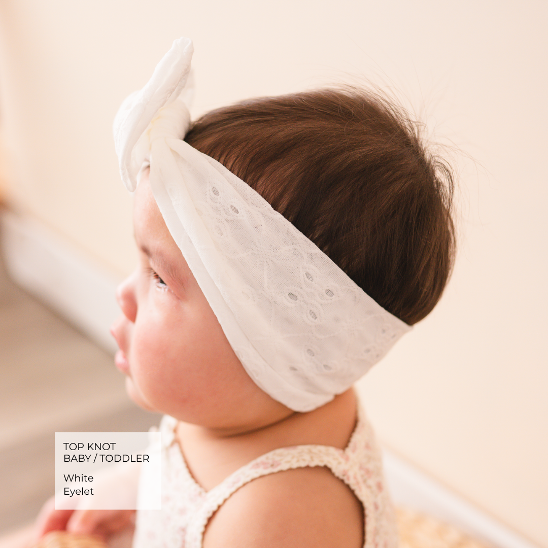 A baby wearing a white eyelet top-knot headband, shown in profile against a soft cream background. A text overlay in the bottom-left corner reads 'TOP KNOT BAB / TODDLER White Eyelet'.
