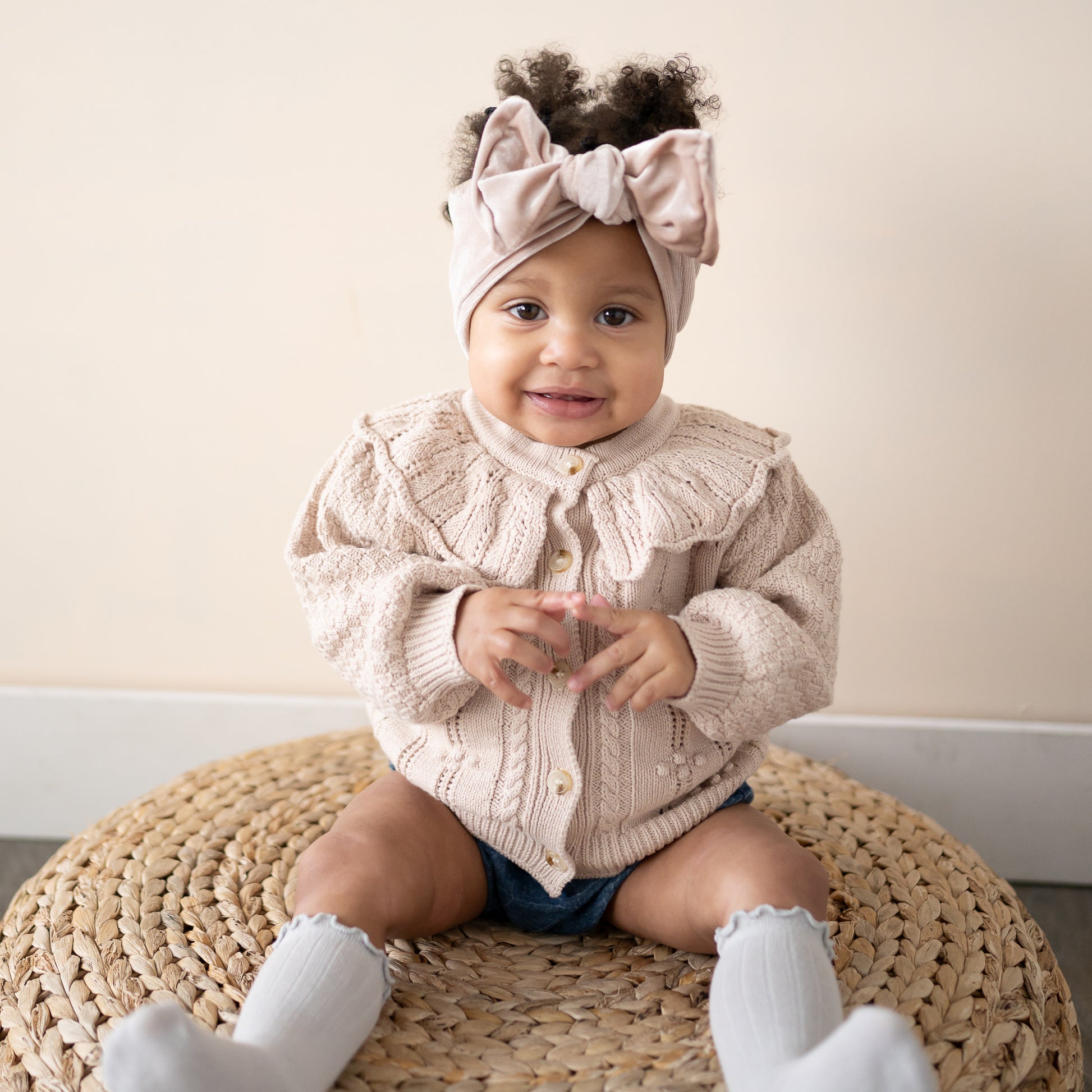 Baby wearing a beige knitted cardigan and headband sitting on a woven mat.