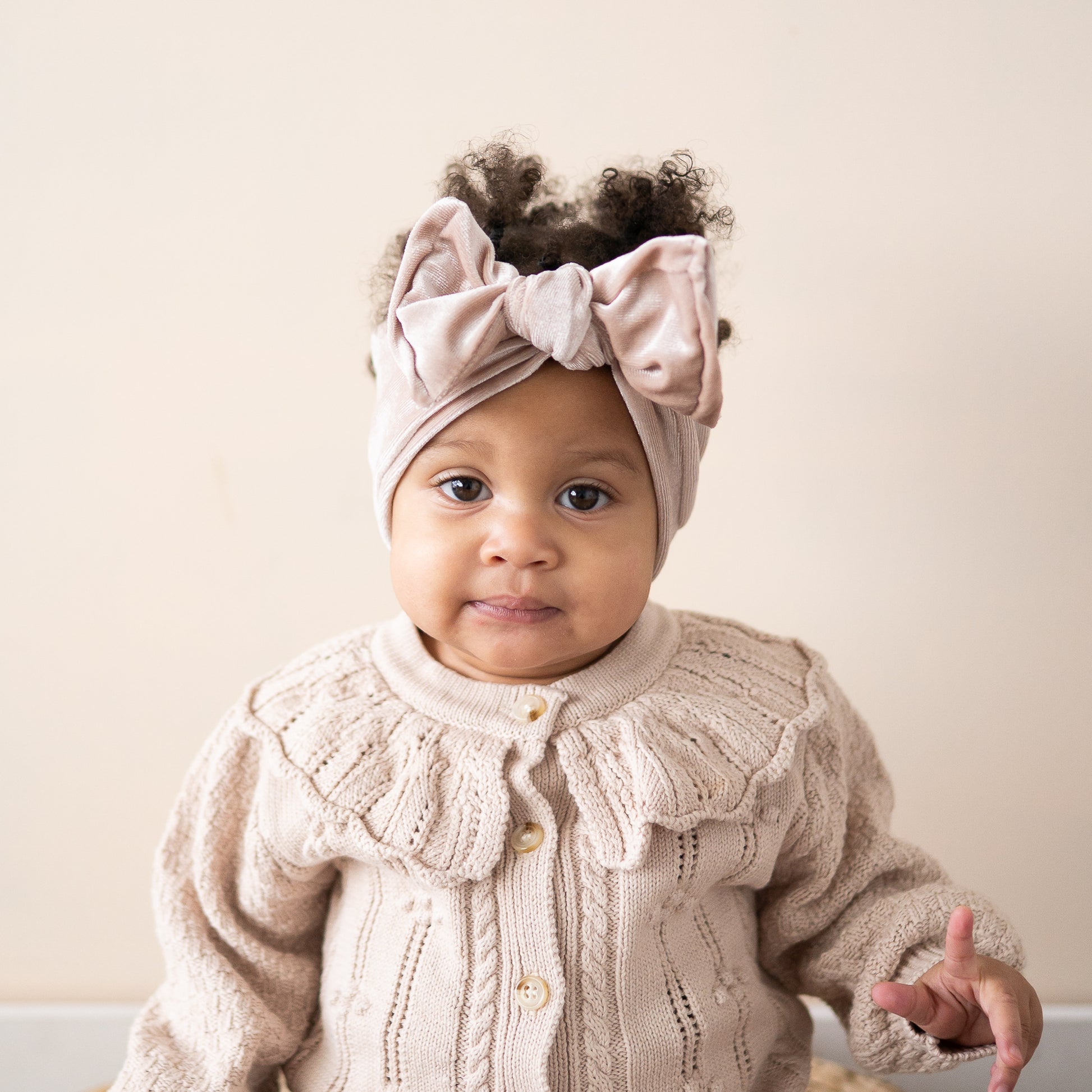 Baby wearing a beige knit cardigan and velvet headband against a plain background