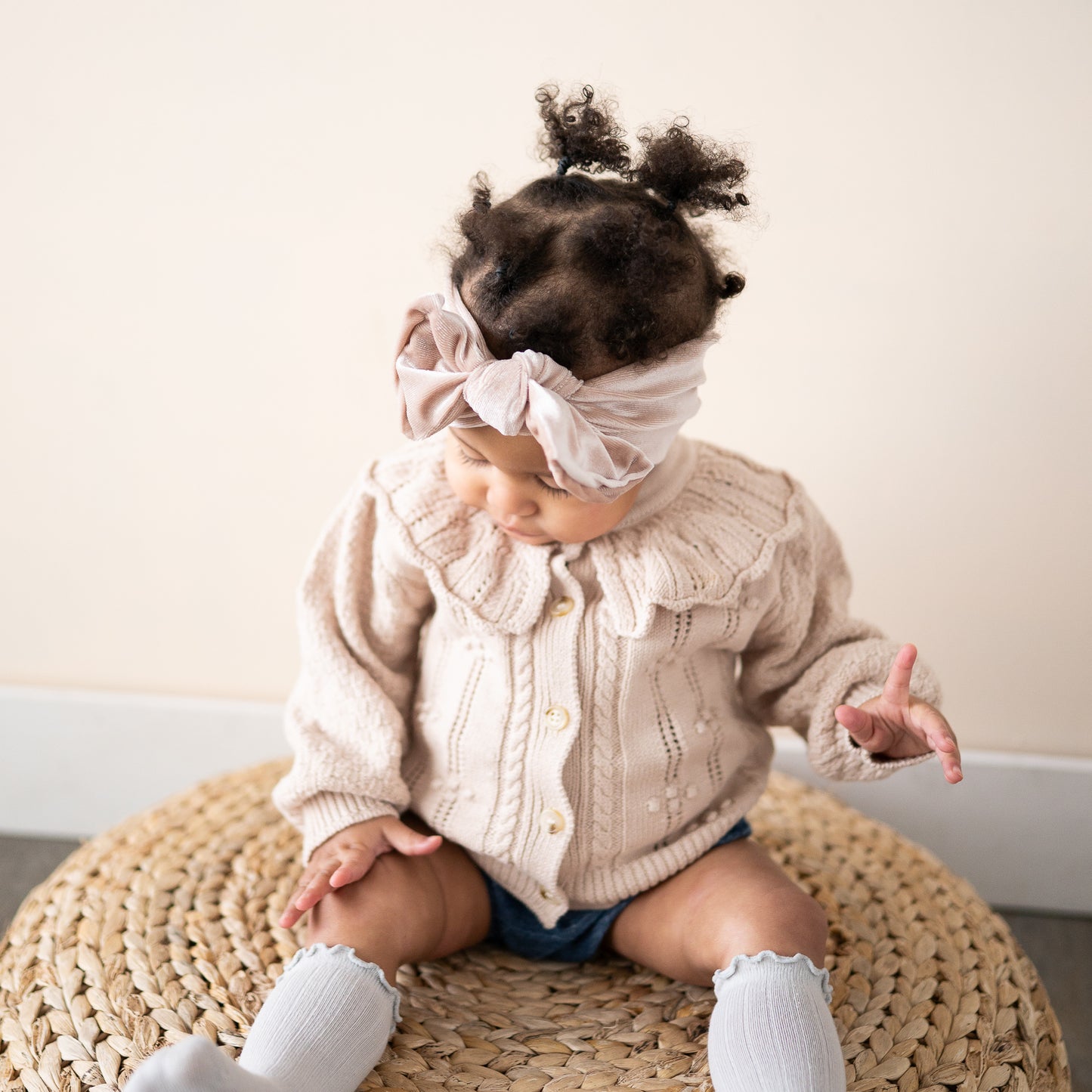 Baby wearing a neutral velvet top knot headband, beige cardigan and white socks sitting on a woven stool.