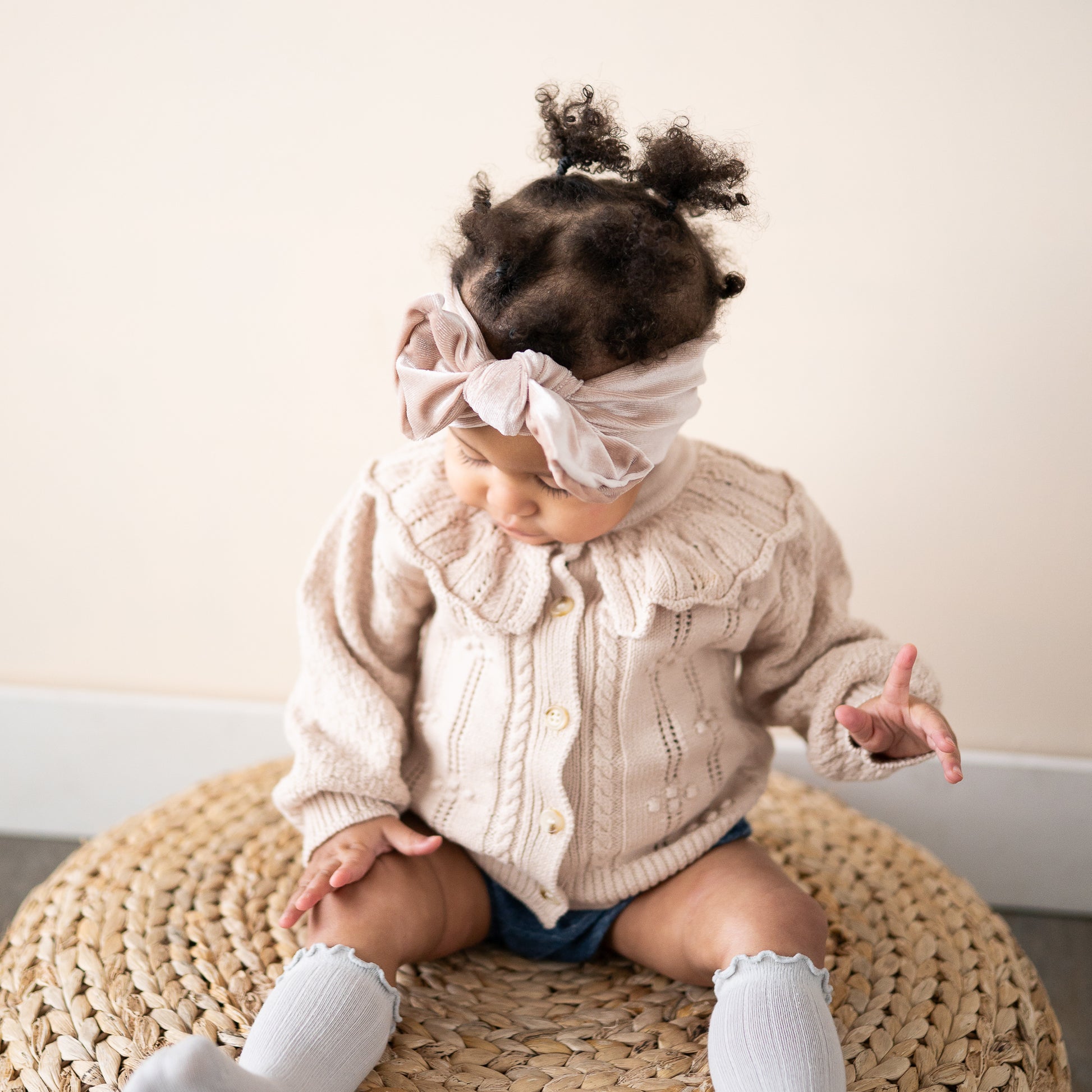 Baby wearing a neutral velvet top knot headband, beige cardigan and white socks sitting on a woven stool.