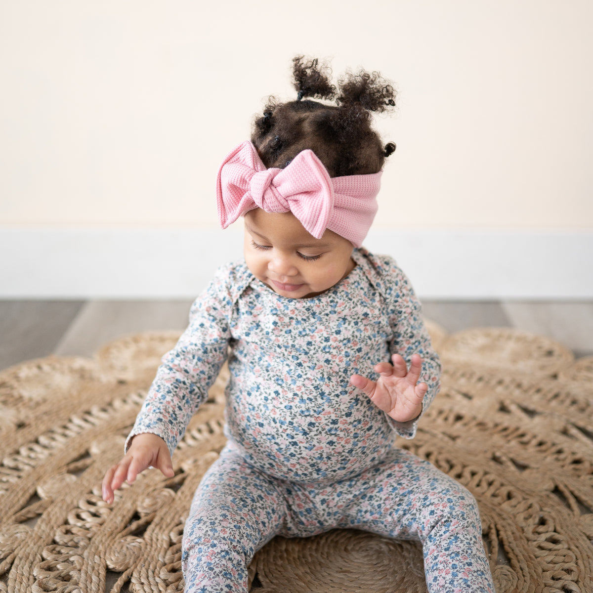 Baby wearing a floral outfit and pink headband sitting on a textured rug.