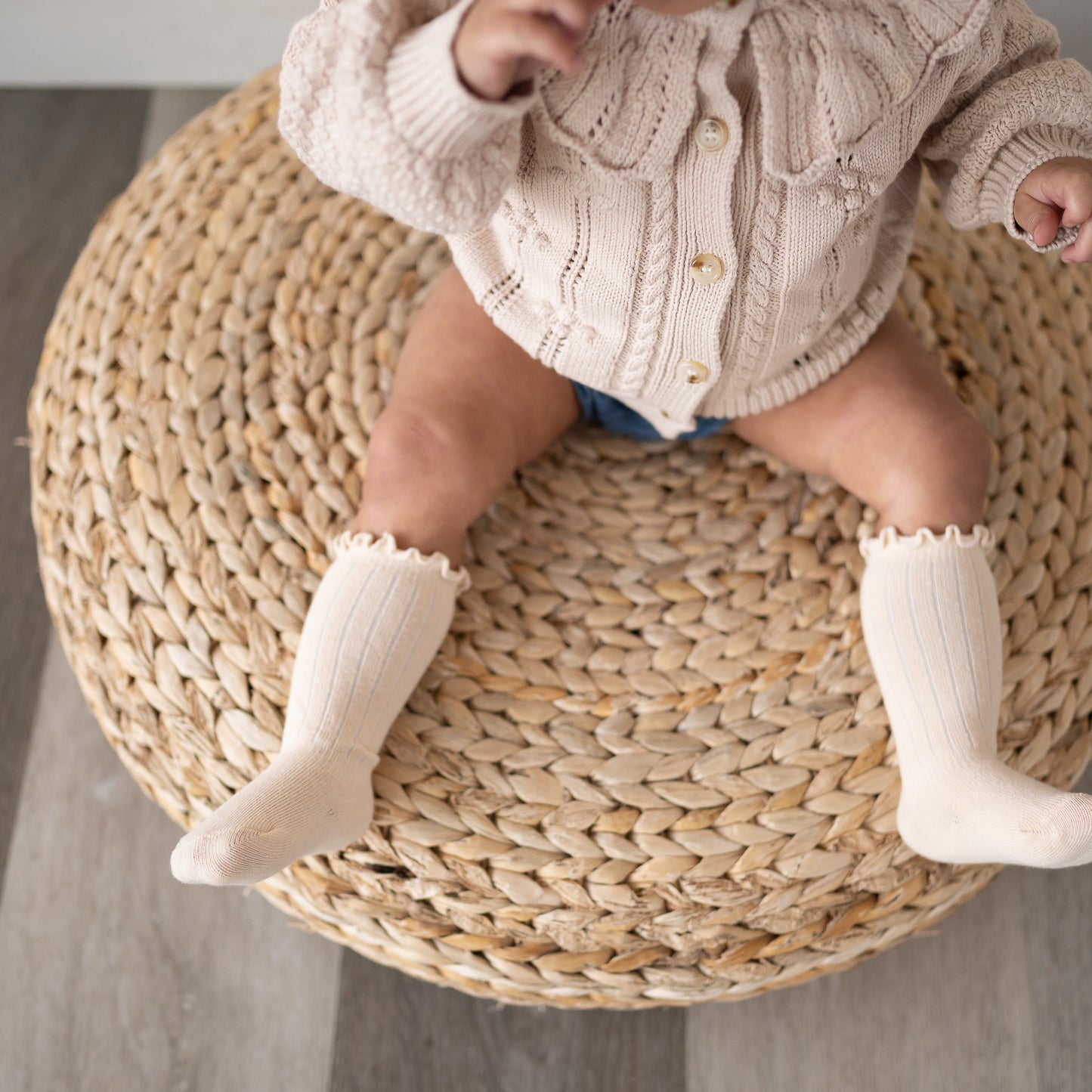 Baby sitting on a woven stool wearing a knitted cardigan and white socks.