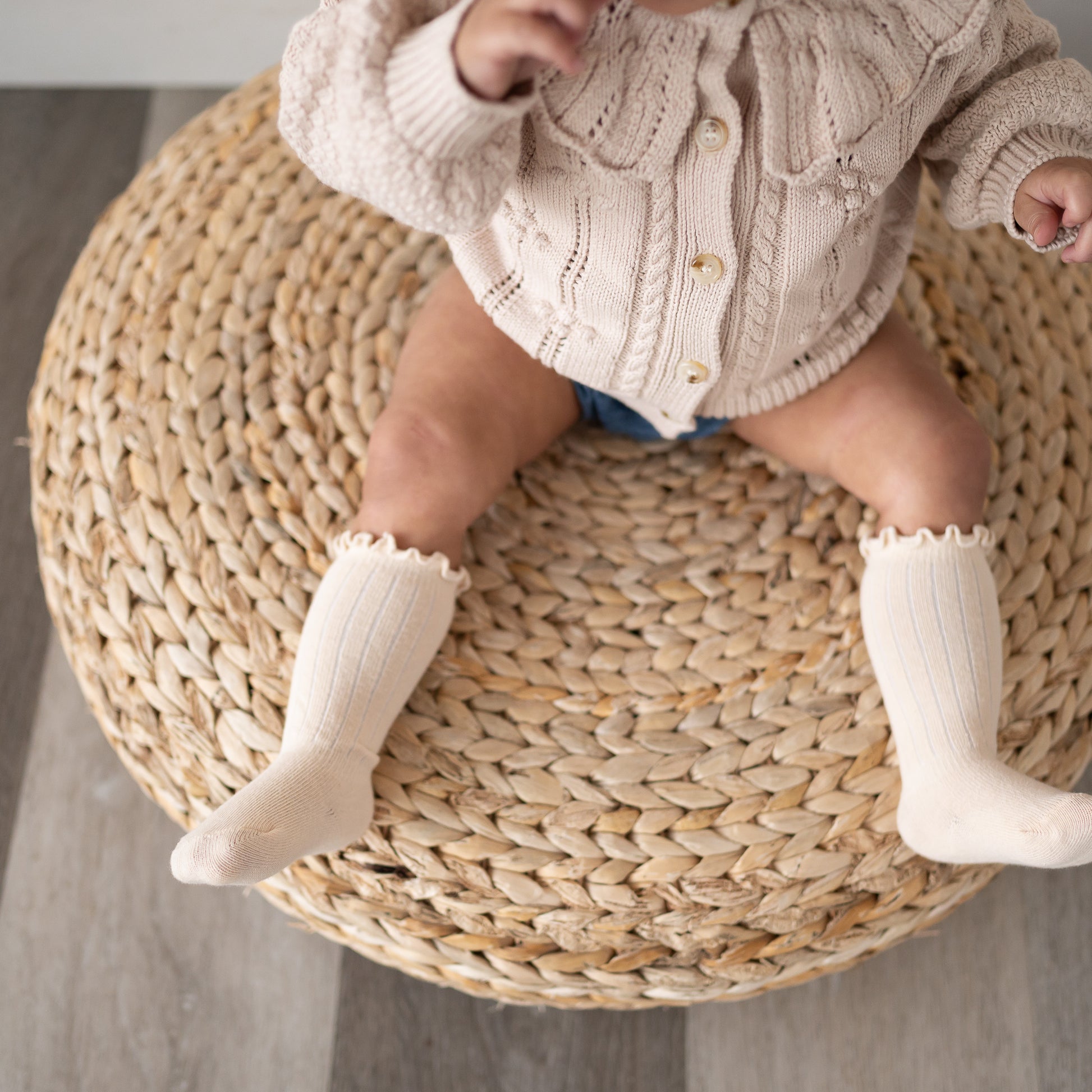 Baby sitting on a woven stool wearing a knitted cardigan and white socks.