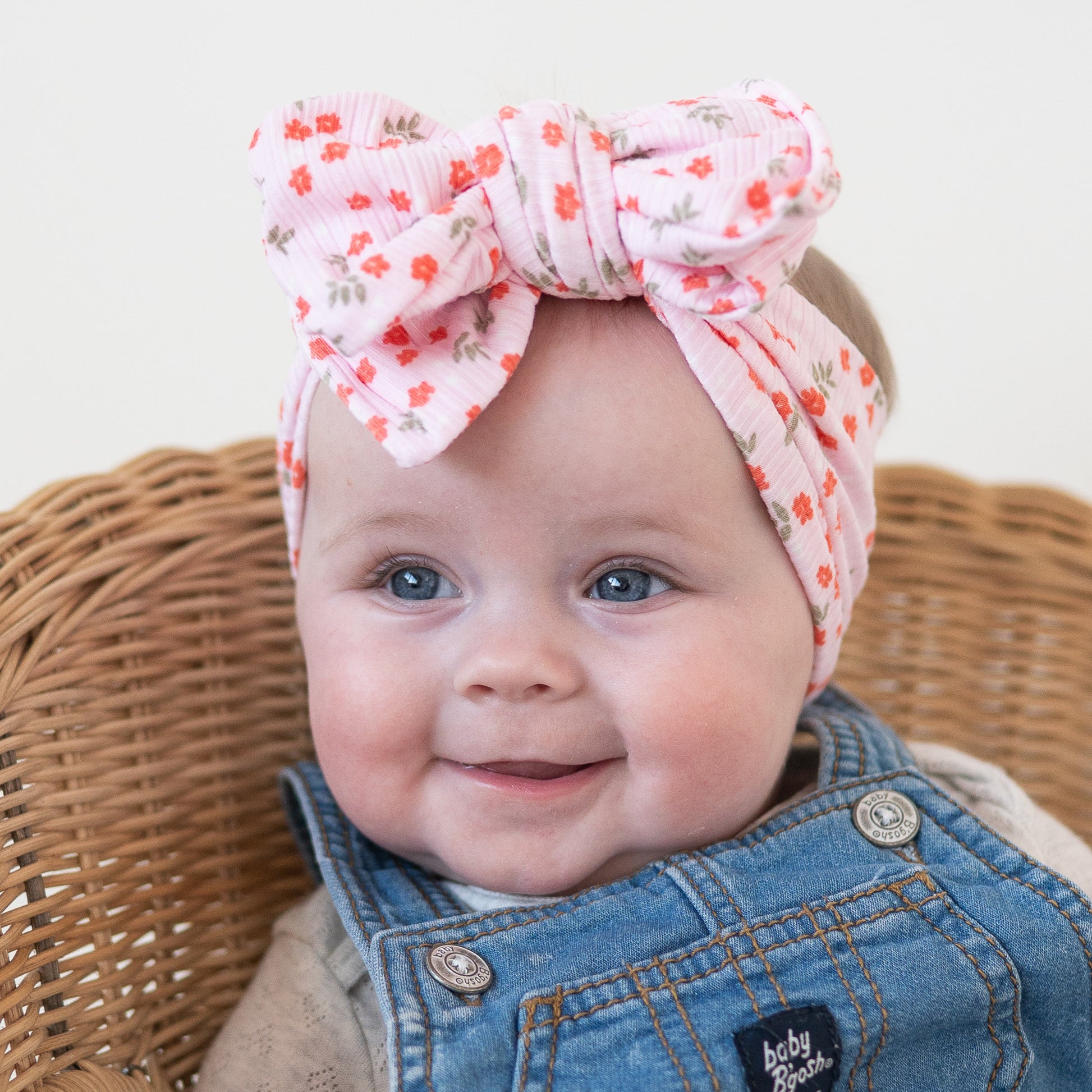 Baby wearing a floral headband and denim outfit sitting in a wicker chair.
